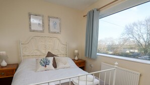 A bedroom with a white metal bed frame, a wooden nightstand with a lamp, a window with blue curtains, and a view of trees and buildings outside. - Towyn Hall (New Quay)