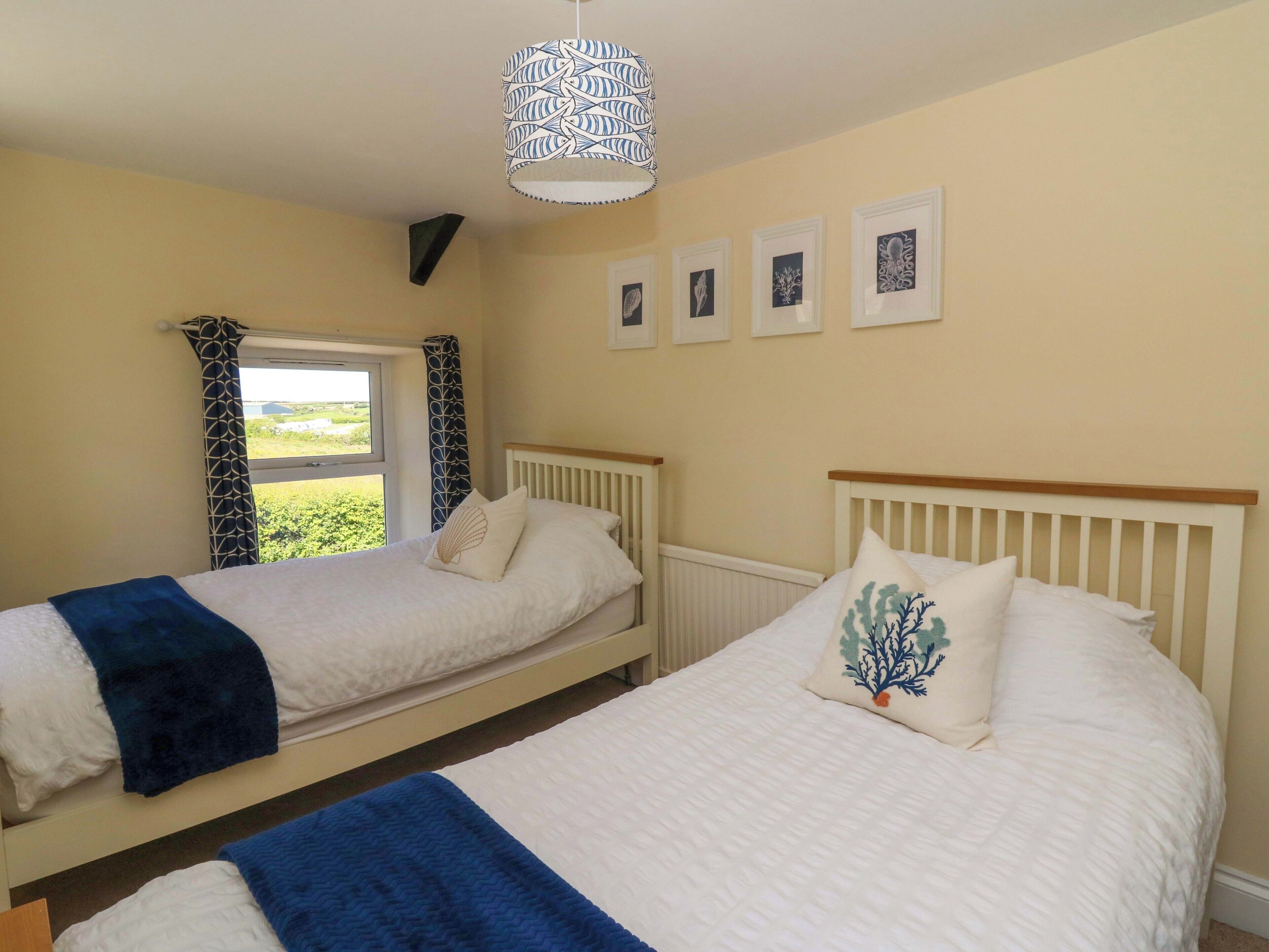 A bedroom with two single beds, a window with curtains, a hanging light fixture, and framed pictures on the wall.