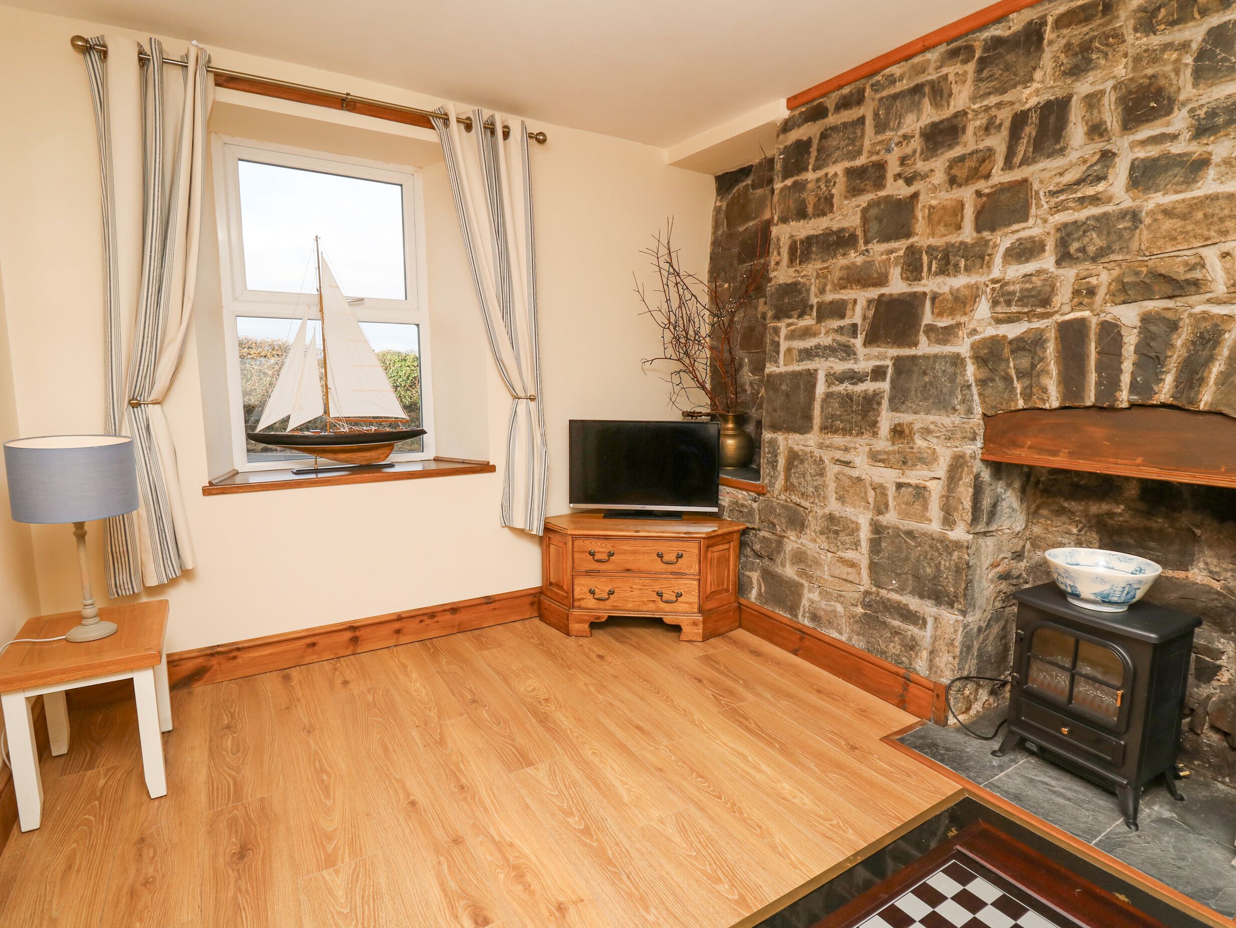 A cozy room with a wooden floor, a stone fireplace, a television on a wooden cabinet, and a window with curtains.