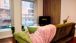 Un salón con un sofá verde, una manta rosa, un televisor mostrando 'NETFLIX' y una ventana con persianas.