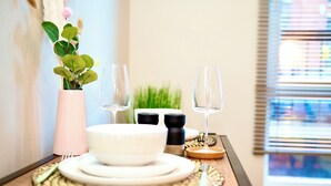 Una mesa de comedor dispuesta con platos, tazones, cubiertos y vasos, junto a una jarra con follaje verde y saleros y pimenteros.