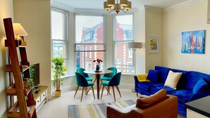 A modern living room with a blue sofa, a wooden TV stand, a dining table with chairs, and large windows with blinds. - Stylish Apartment with views, lift and parking (Scarborough)