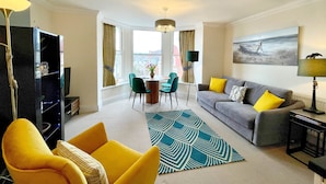 Un salon moderne avec un canapé gris, des coussins jaunes, un tapis à motifs bleus et un coin repas avec des chaises vertes.