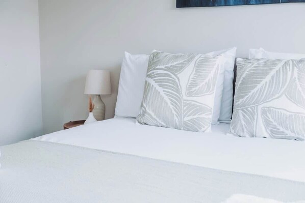 A bed with white bedding and leaf-patterned pillows, a bedside table with a lamp, and a wall-mounted artwork.