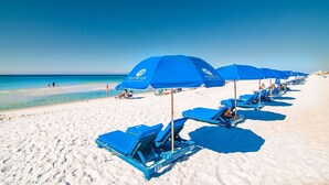 Una playa con hileras de sombrillas azules y tumbonas, un cielo despejado y el océano al fondo.
