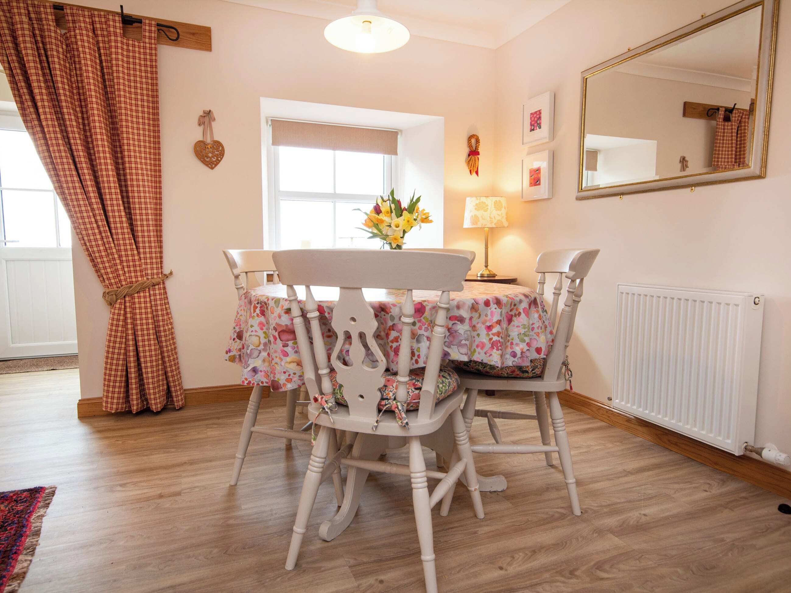 A dining area with a table set for four, surrounded by white chairs, a window with a view, a lamp, and a radiator.