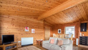 Ein gemütliches Blockhaus-Interieur mit Holzbalkendecke, einem Fernseher, einer Couch und einer Essecke mit Stühlen.