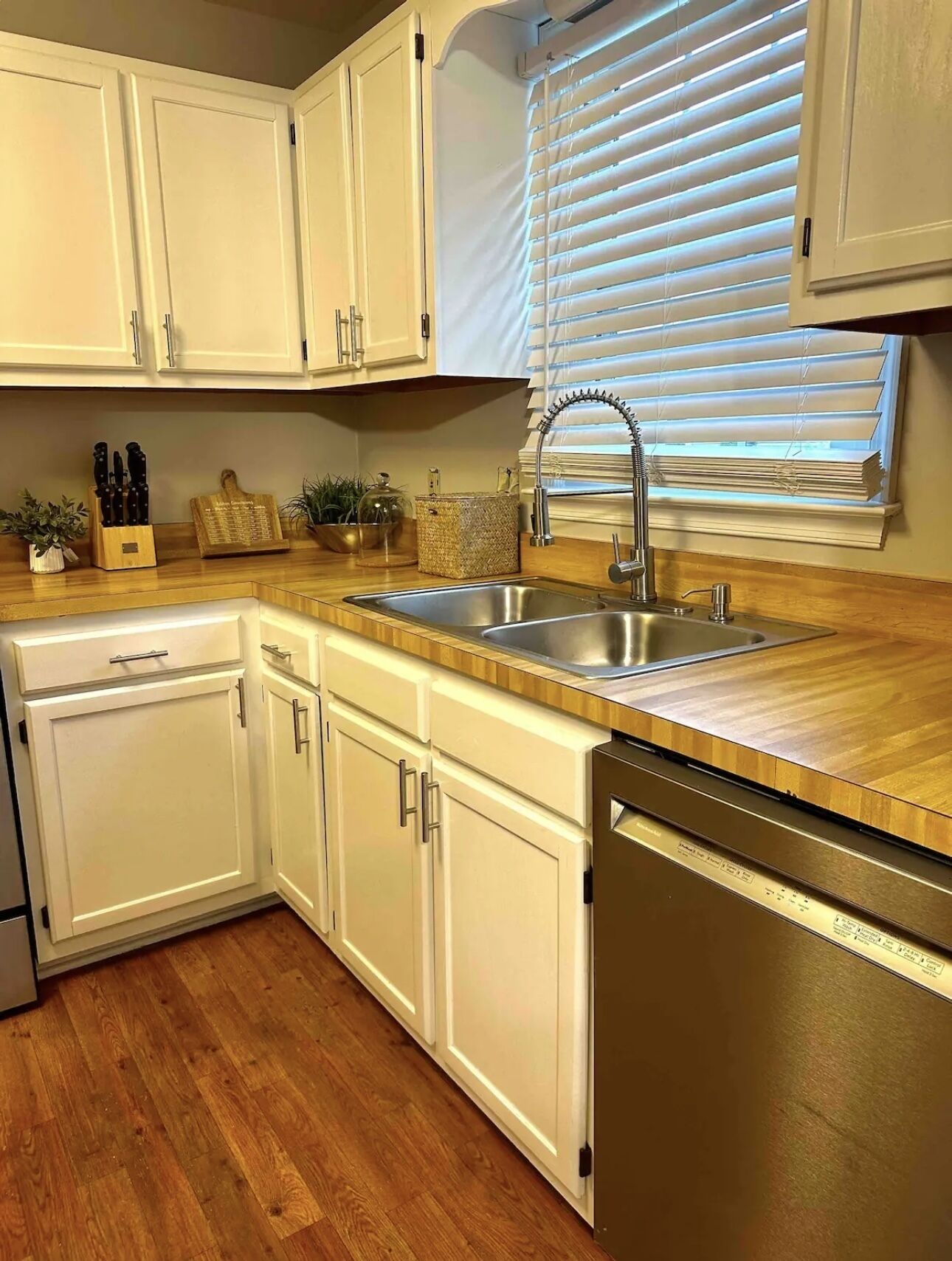 A kitchen with white cabinets, a stainless steel sink, wooden countertops, and a dishwasher.