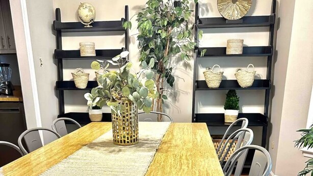 A dining area with a wooden table, grey chairs, a woven table runner, and shelves with decorative items and a plant.