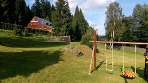En græsplæne med volleyballnet, gyngestativ og en rød bygning i baggrunden.