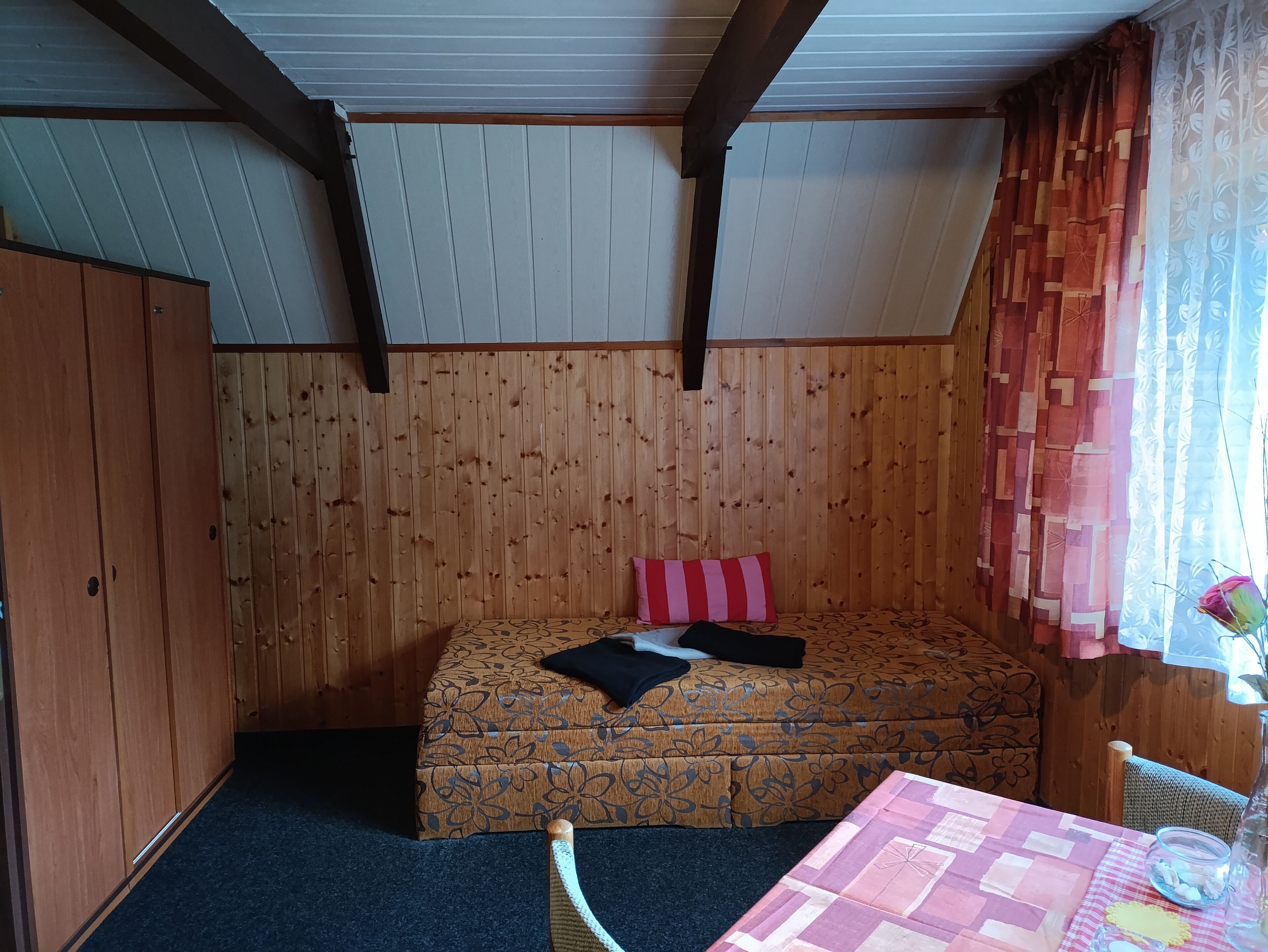 A room with a wooden paneled wall, a bed with a floral bedspread, a small table with a patterned tablecloth, and a window with patterned curtains.
