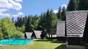 Et poolområde med en blå pool, omgivet af en græsplæne og træer. Der er tre træhytter med spidse tage.