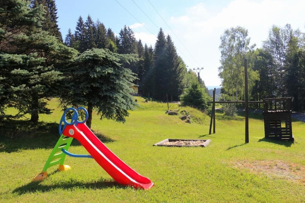 A playground with a red slide and climbing structures in a grassy area with trees and a wooden playhouse in the background.