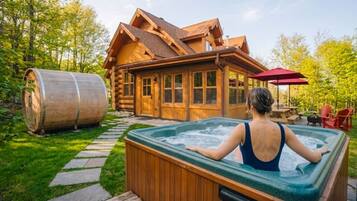 A log cabin with a hot tub in front, surrounded by a wooden deck and a stone pathway.