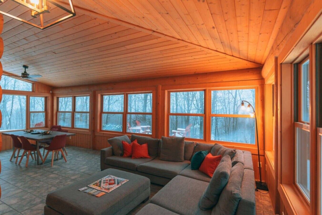 A cozy cabin interior with a wooden ceiling, large windows, a dining table, and a sofa.