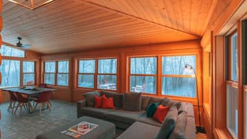 A cozy cabin interior with a wooden ceiling, large windows, a dining table, and a sofa.