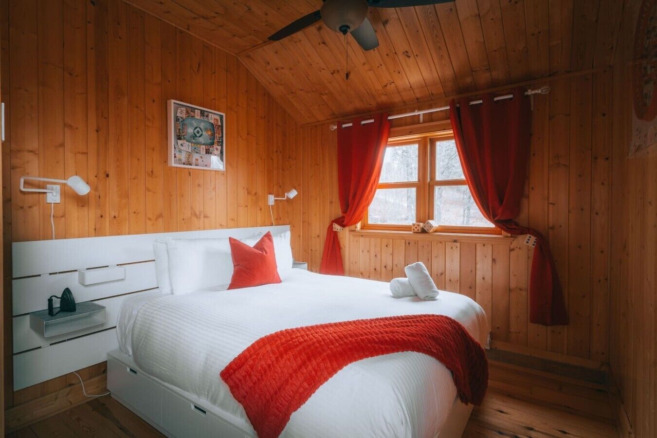 A wooden cabin bedroom with a bed, white headboard, red curtains, and a window.