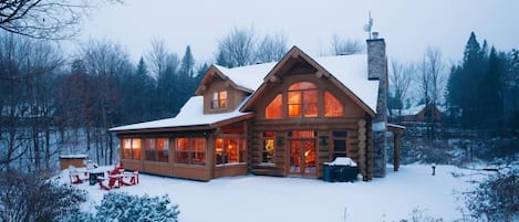 Uma cabana de tronco com um alpendre grande e várias janelas, rodeada por uma paisagem coberta de neve.