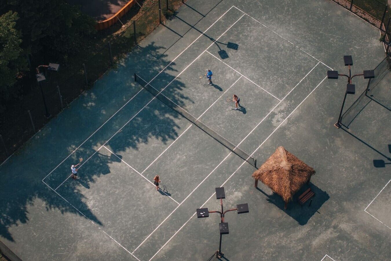 An aerial view of a tennis court with a thatched-roof structure, surrounded by trees and equipped with multiple light poles.