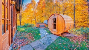 Una sauna de barril de madera en un bosque, con un sendero de piedra que lleva a un edificio de madera.