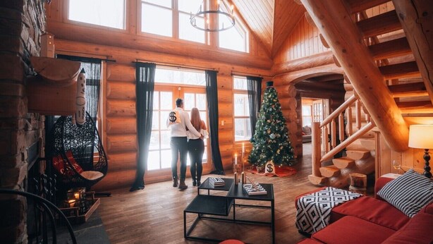 Interior de una cabaña de troncos con una escalera de madera, un árbol de Navidad, un sofá y una mesita de centro.