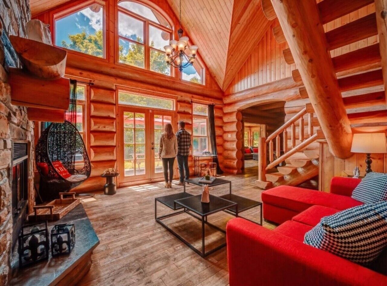 Interior de uma cabana de madeira com lareira, uma cadeira suspensa, escada e um sofá vermelho.