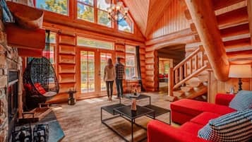 Interior de uma cabana de madeira com lareira, uma cadeira suspensa, escada e um sofá vermelho.