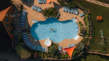 Vista aérea de uma piscina cercada por espreguiçadeiras e uma banheira de hidromassagem, com um edifício e vegetação ao fundo.