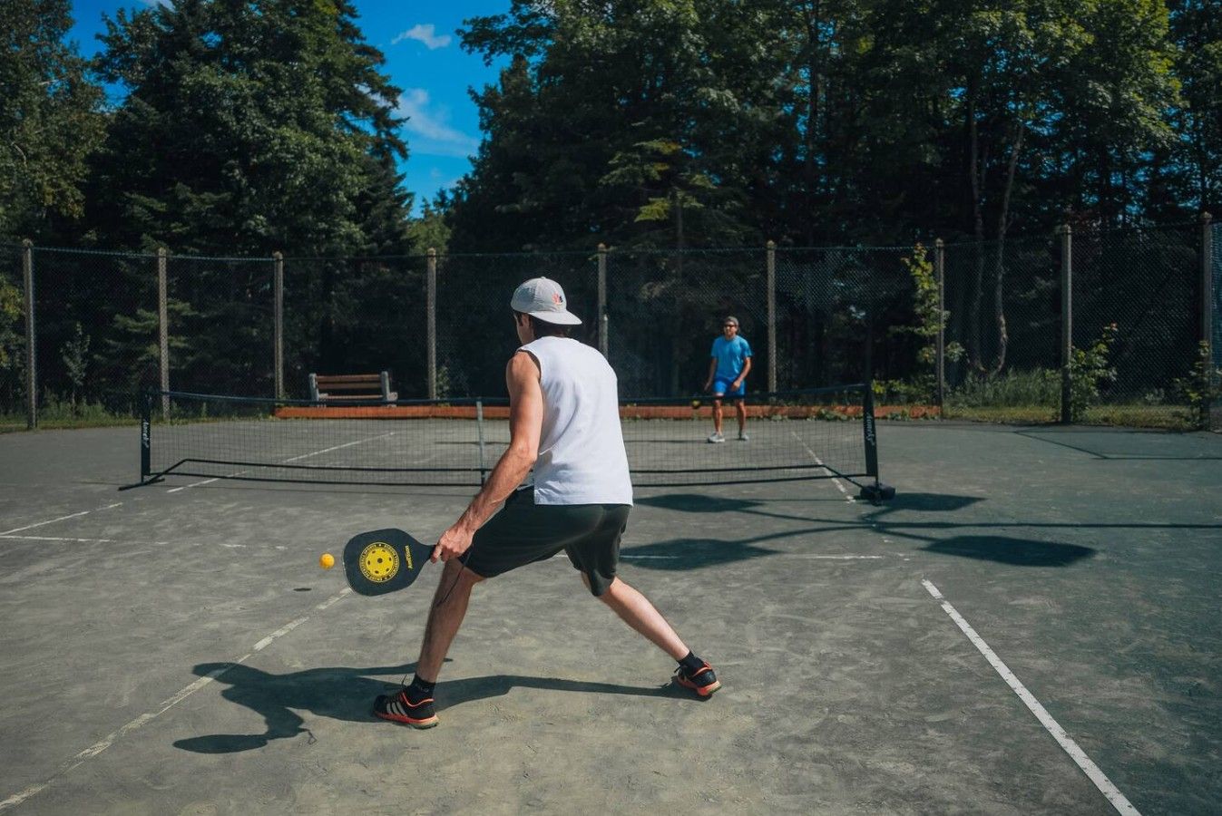 Una cancha de tenis al aire libre con red, rodeada por una valla y árboles.