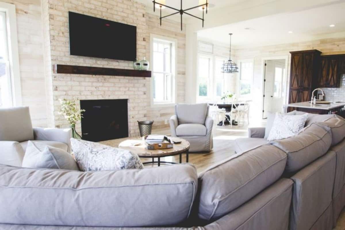Una sala de estar moderna con un sofá grande de color gris, una chimenea, un televisor de pantalla plana montado en una pared de ladrillo, y un comedor visible a través de una puerta abierta.