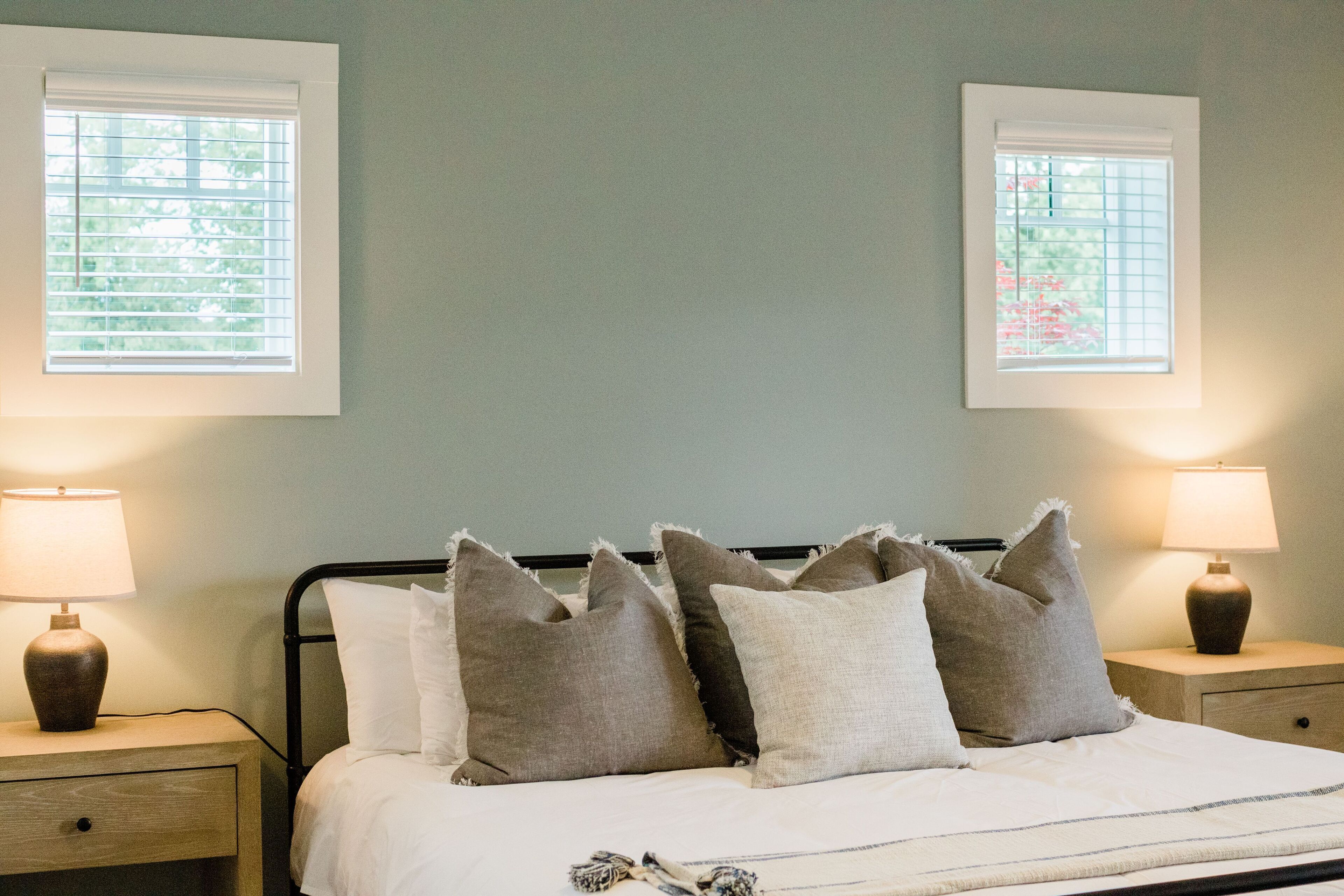 A neatly made bed with white and gray pillows, two bedside lamps, and a window with blinds.
