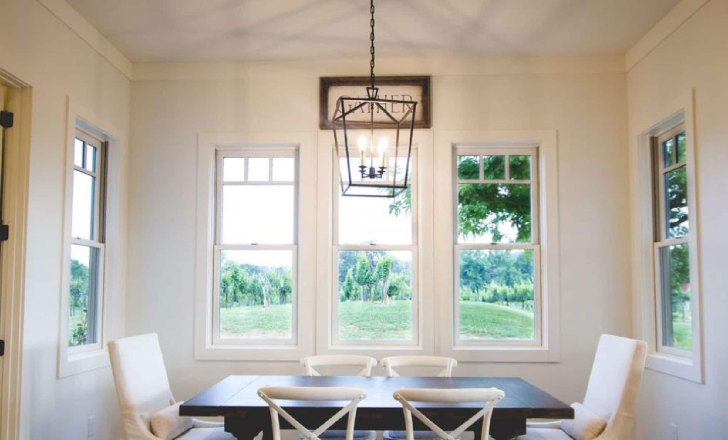 A dining room with a rectangular table, four chairs, and three windows offering a view of greenery.