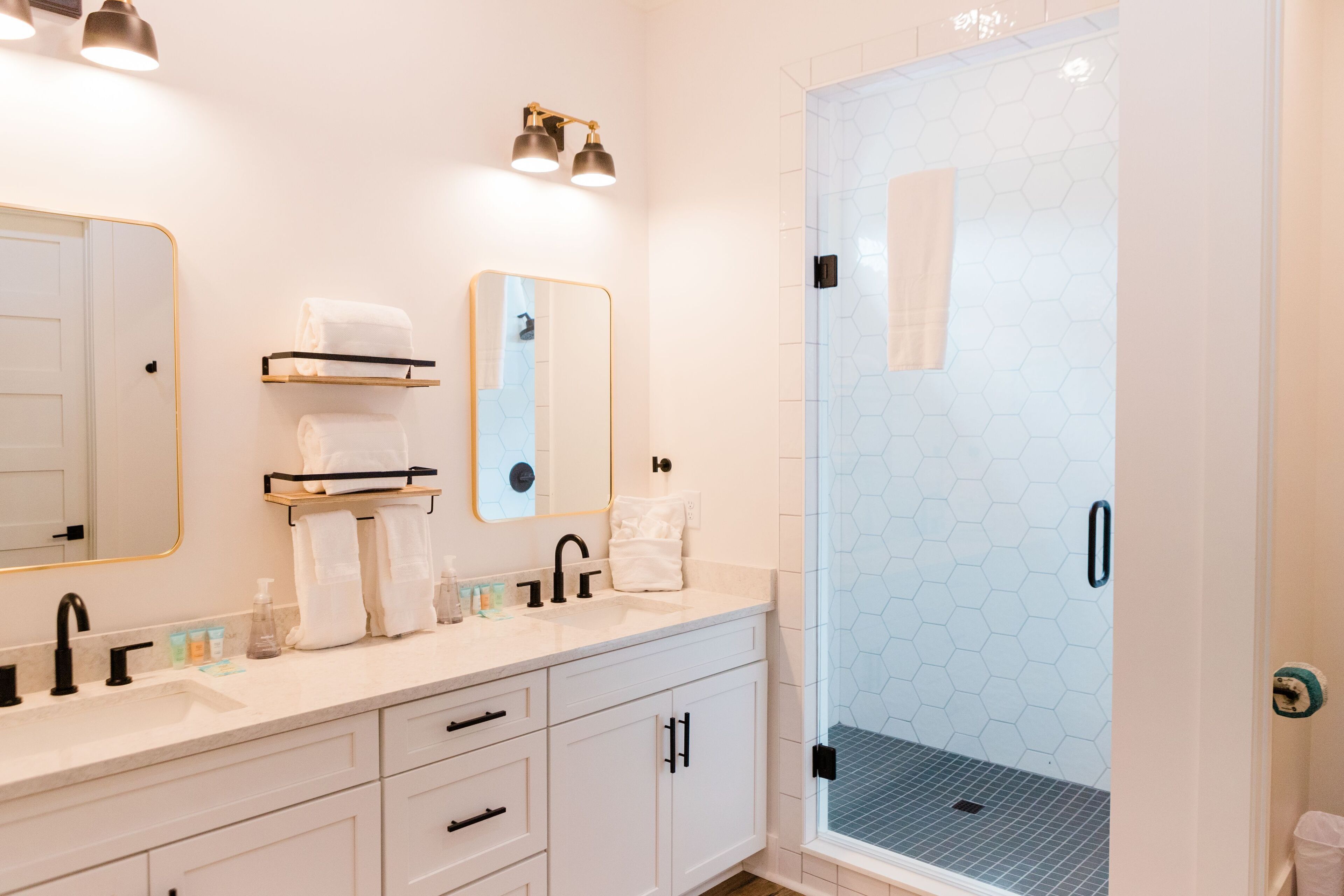 A modern bathroom with a white vanity, two mirrors, a glass shower enclosure, and a hexagonal tile backsplash.