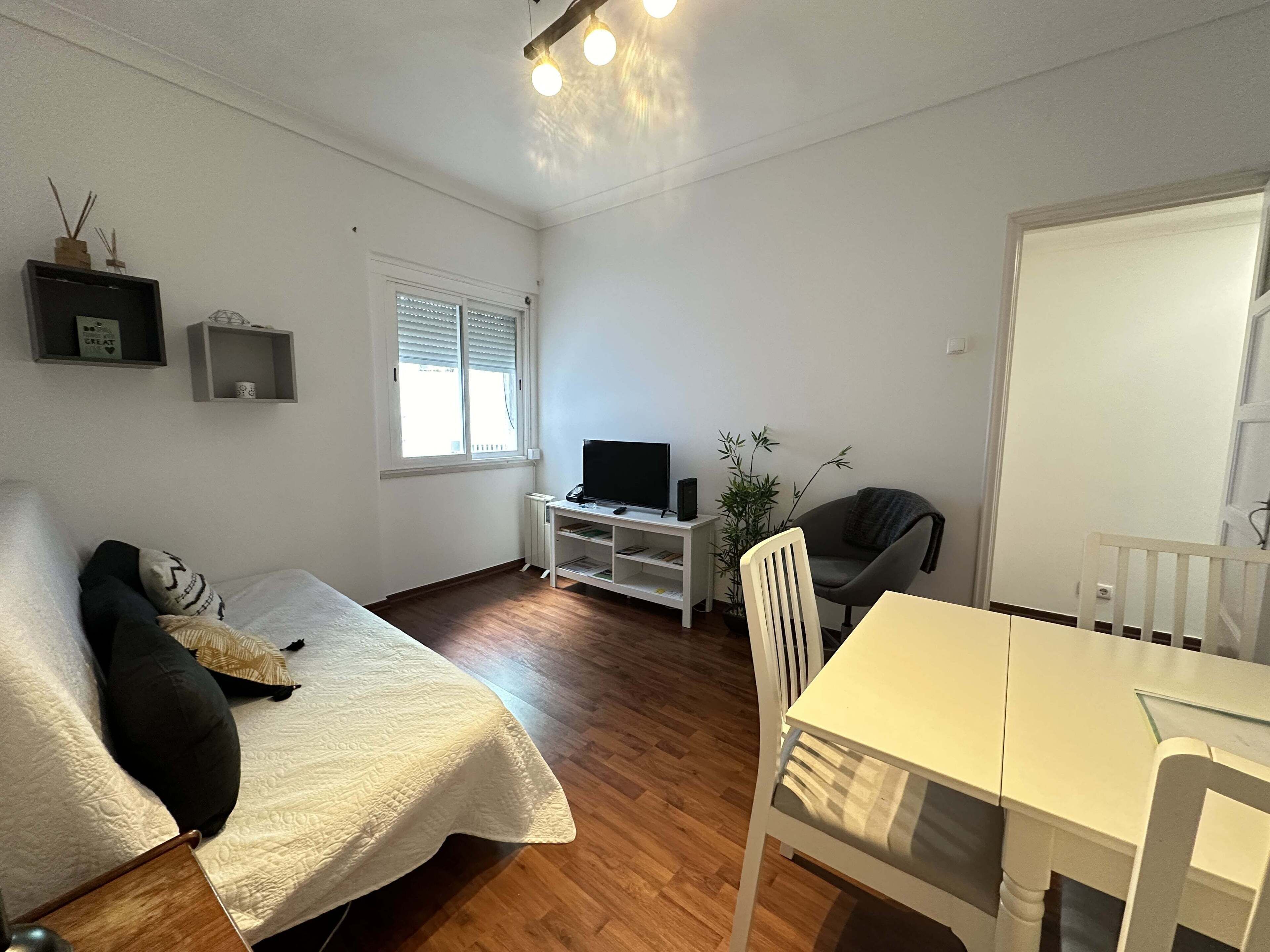 A small, well-lit room with a bed, a TV, a table, and a chair.
