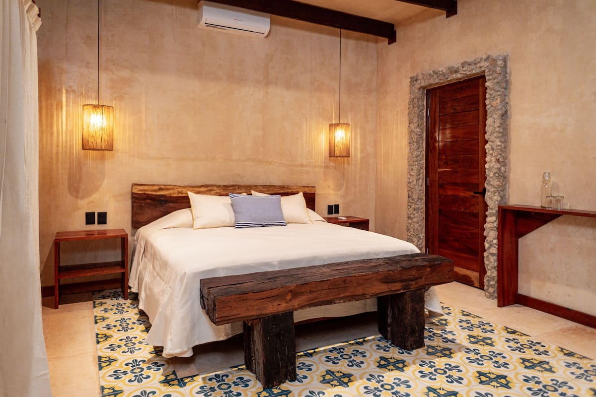 Une chambre avec un lit en bois, deux tables de chevet, un banc, un sol à motifs et une porte encadrée de pierres.