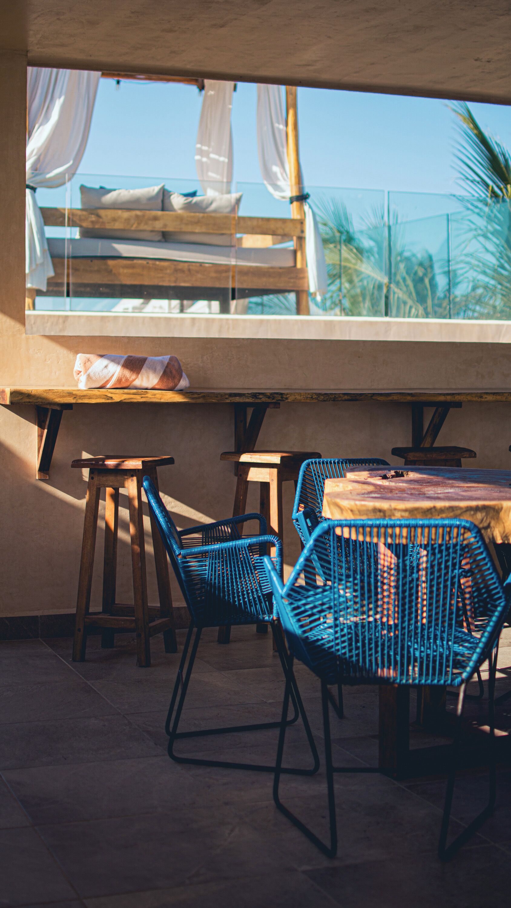 Una terraza con una mesa de madera, sillas azules y vistas a palmeras.