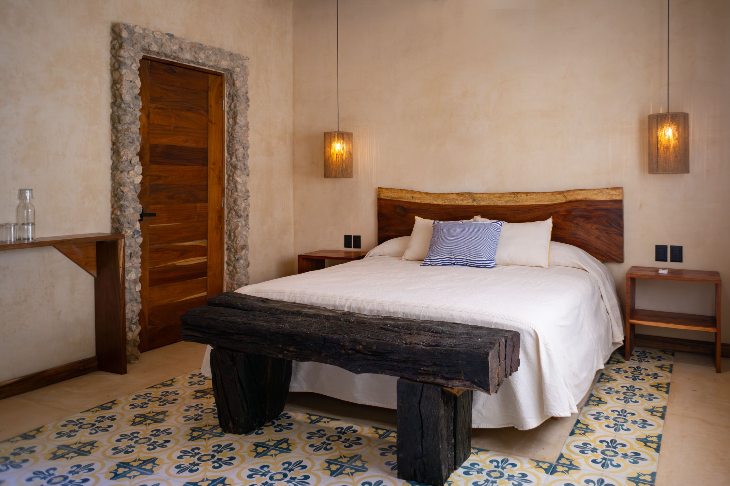 Un dormitorio con una cama de madera, dos mesitas de noche, un banco, una puerta enmarcada en piedra y baldosas de suelo con diseño.