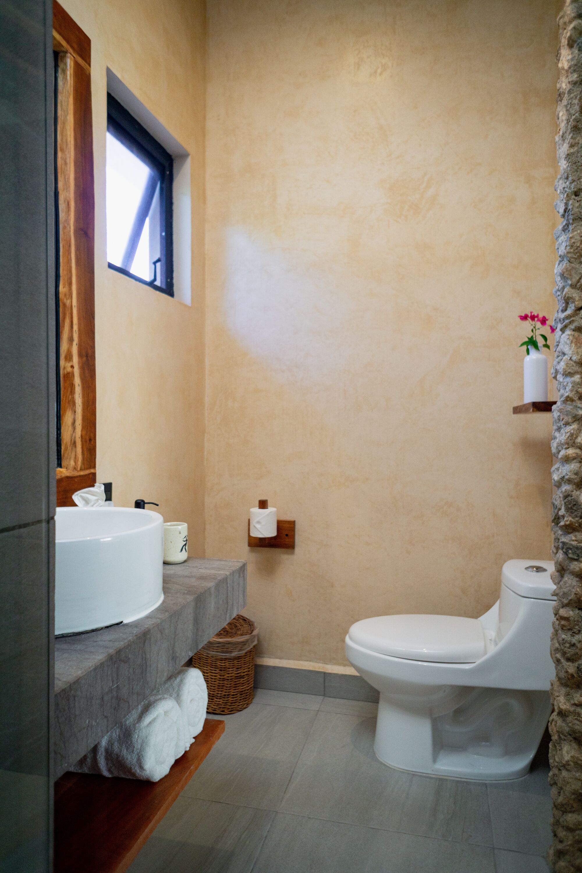 A bathroom with a stone wall, a window, a white sink, a toilet, and a shelf with a vase.