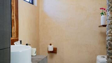 A bathroom with a stone wall, a window, a white sink, a toilet, and a shelf with a vase.
