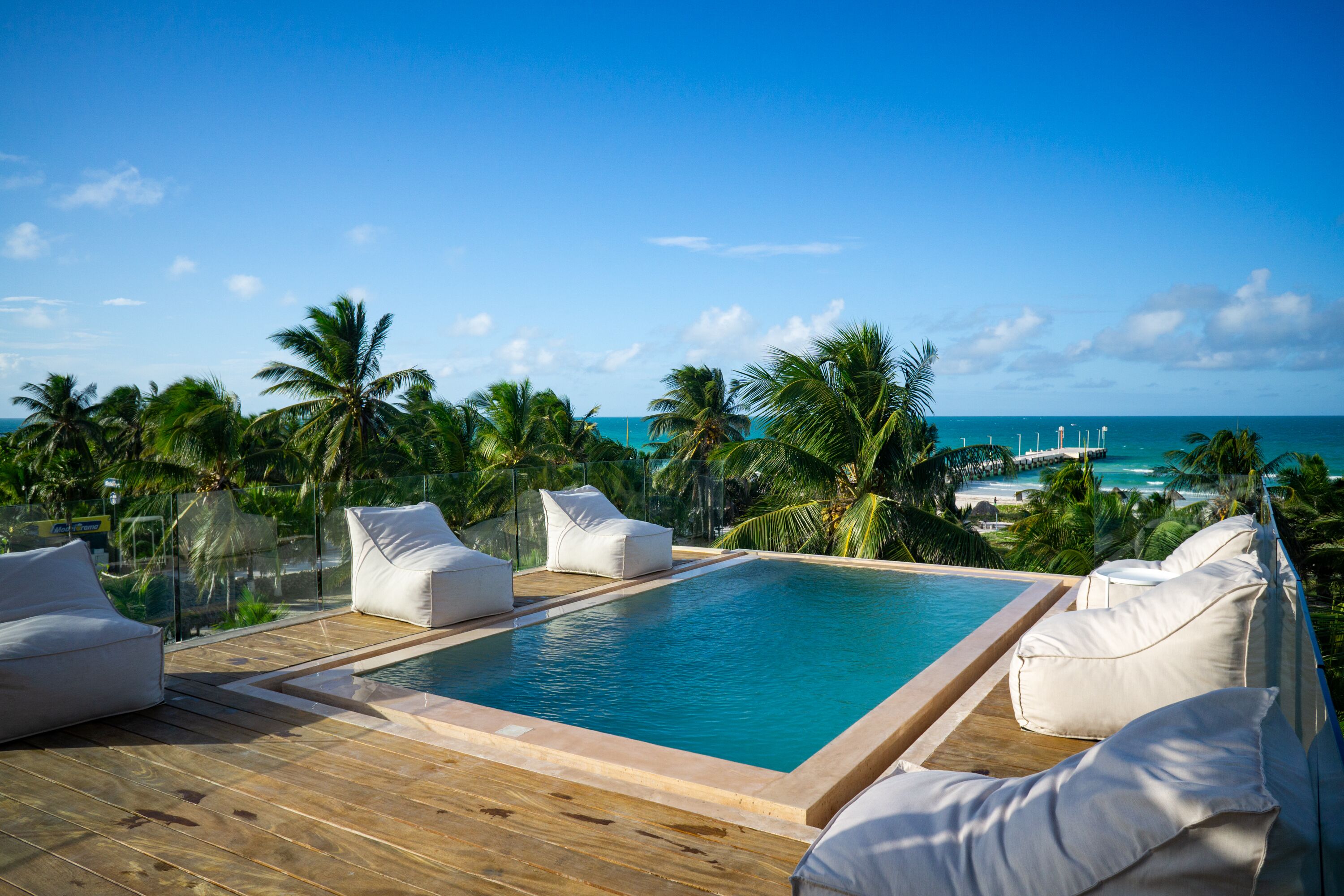 A rooftop infinity pool with white cushioned seating, surrounded by palm trees and overlooking a beach.