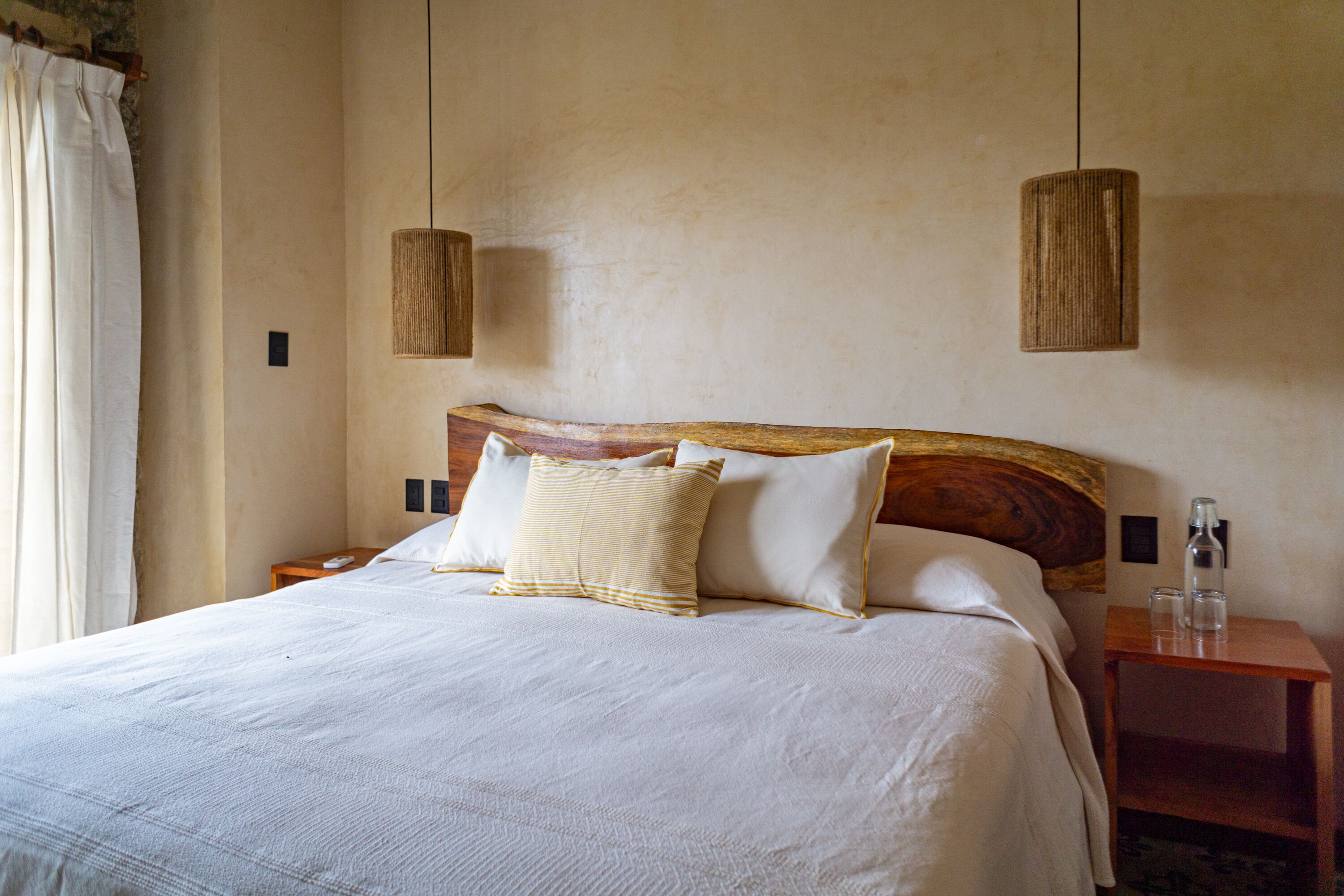 A neatly made bed with white linens and two pillows, a wooden headboard, and two hanging pendant lights.