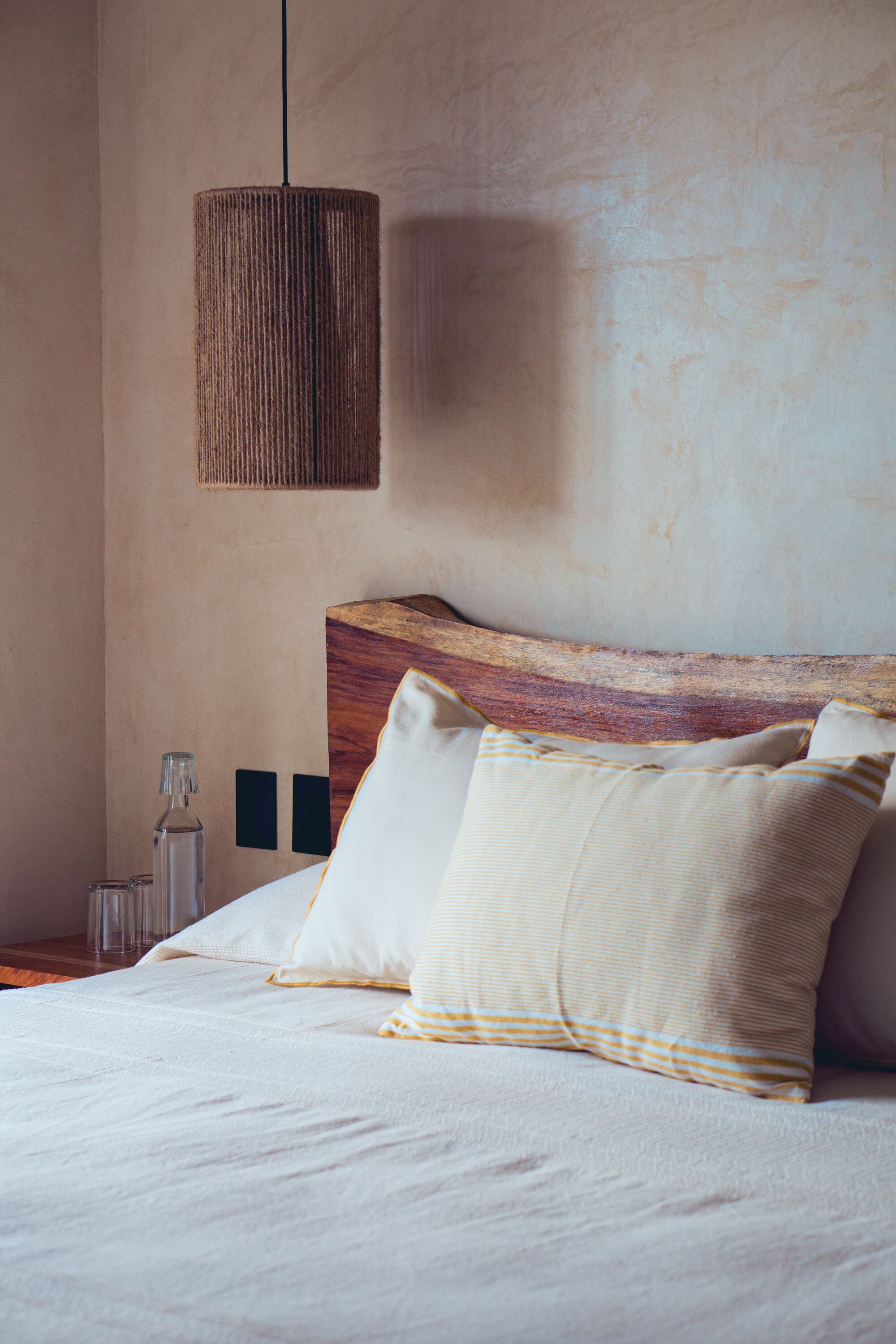 Een slaapkamer met een houten hoofdeinde, een hanglamp en een bed met gestreepte kussens.
