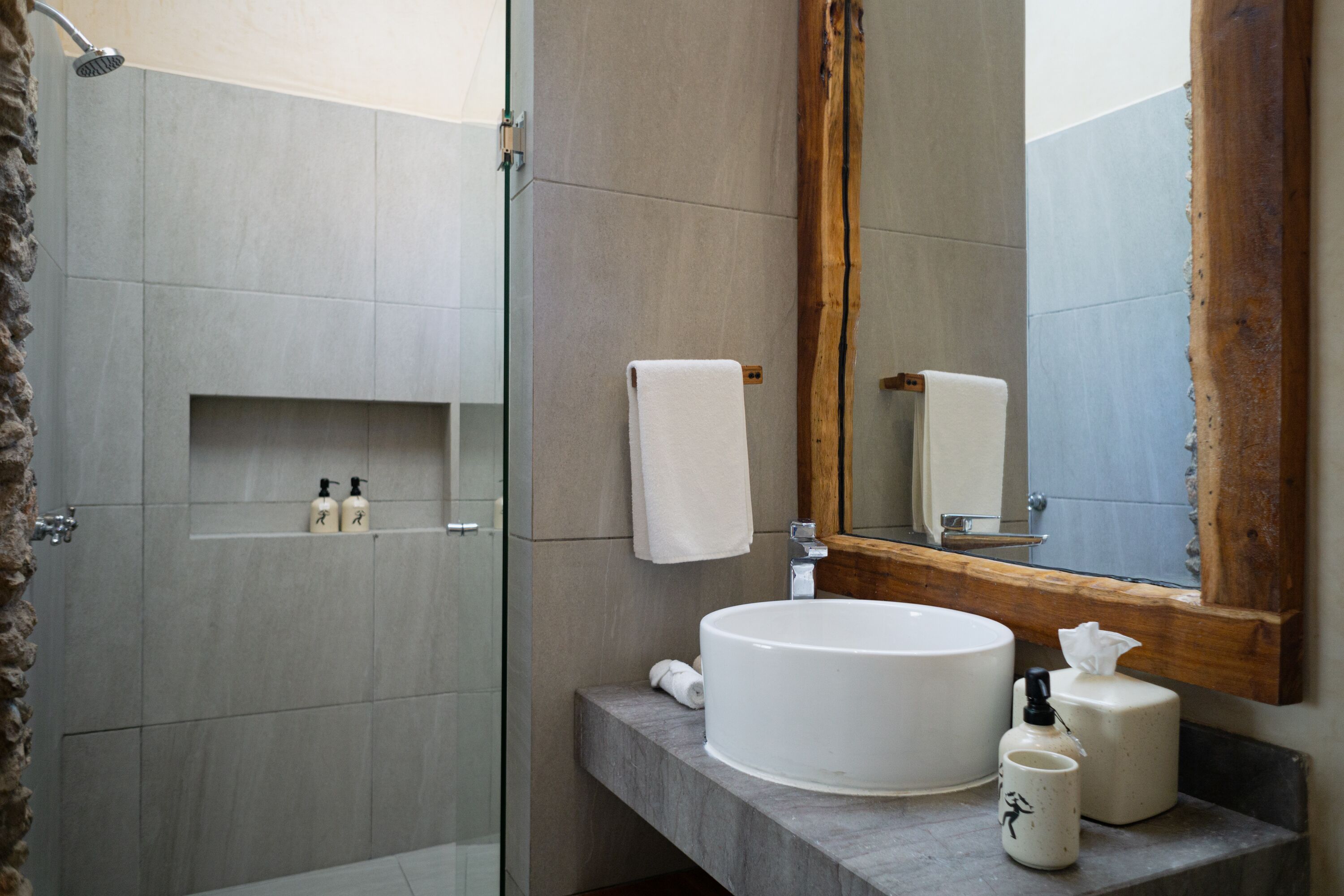 A modern bathroom with a glass shower enclosure, a round sink, and a wooden-framed mirror.