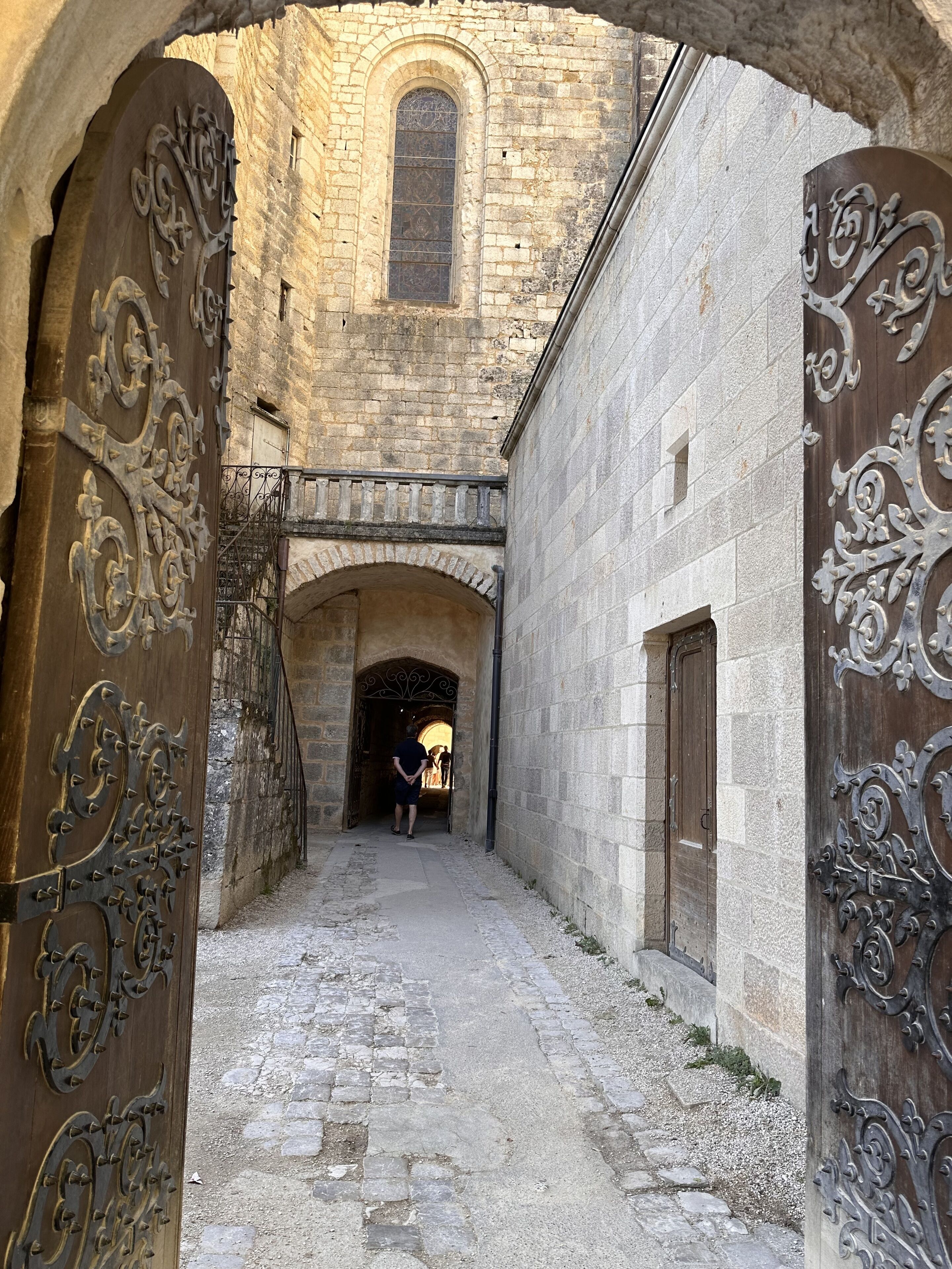 An arched stone corridor with ornate metal doors and a stone building in the background.