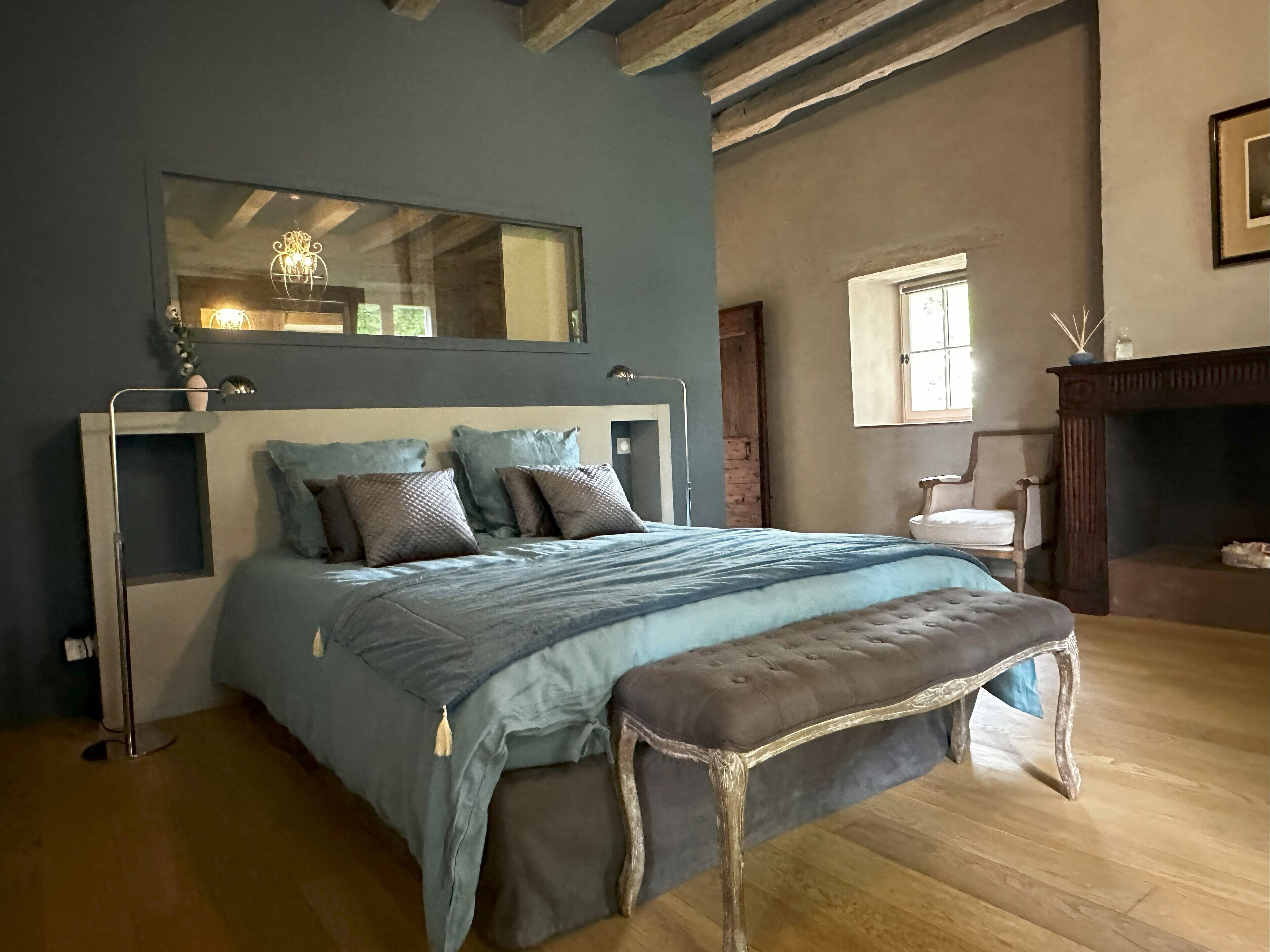 Une chambre à coucher avec un grand lit, un banc en bois, un miroir et une cheminée.