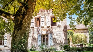 Edifici de pedra amb un gran arbre al davant, una carreta i una zona enjardinada.
