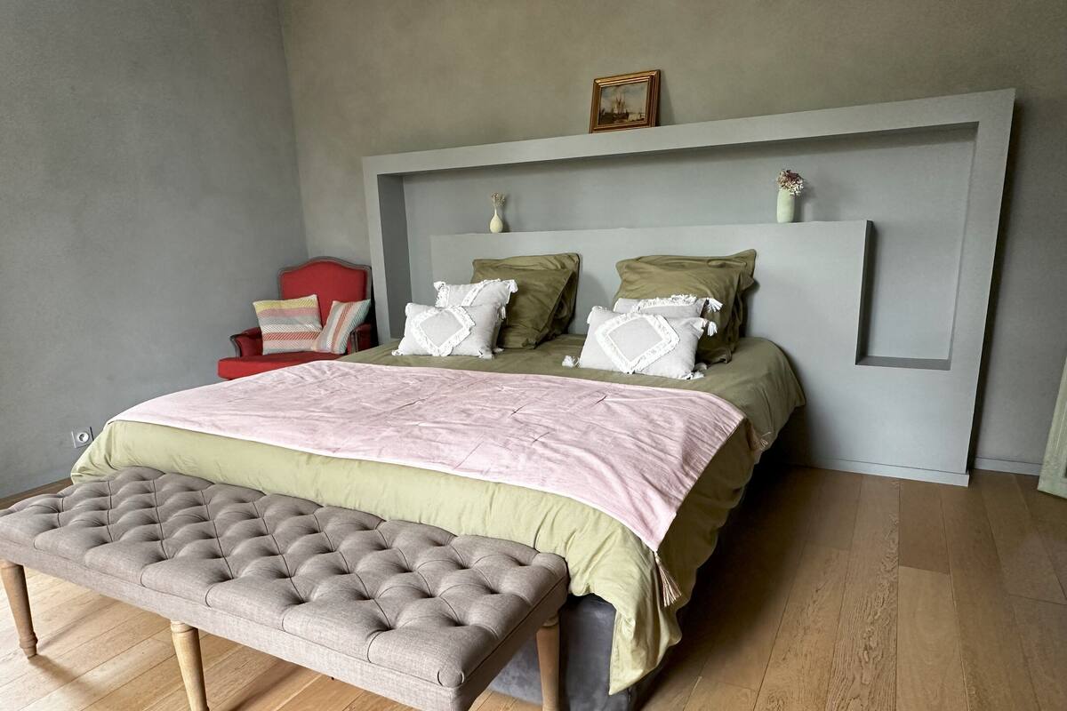 Ein Schlafzimmer mit einem Bett, einer Bank, einem roten Sessel und einem an der Wand befestigten Regal mit einer Vase.