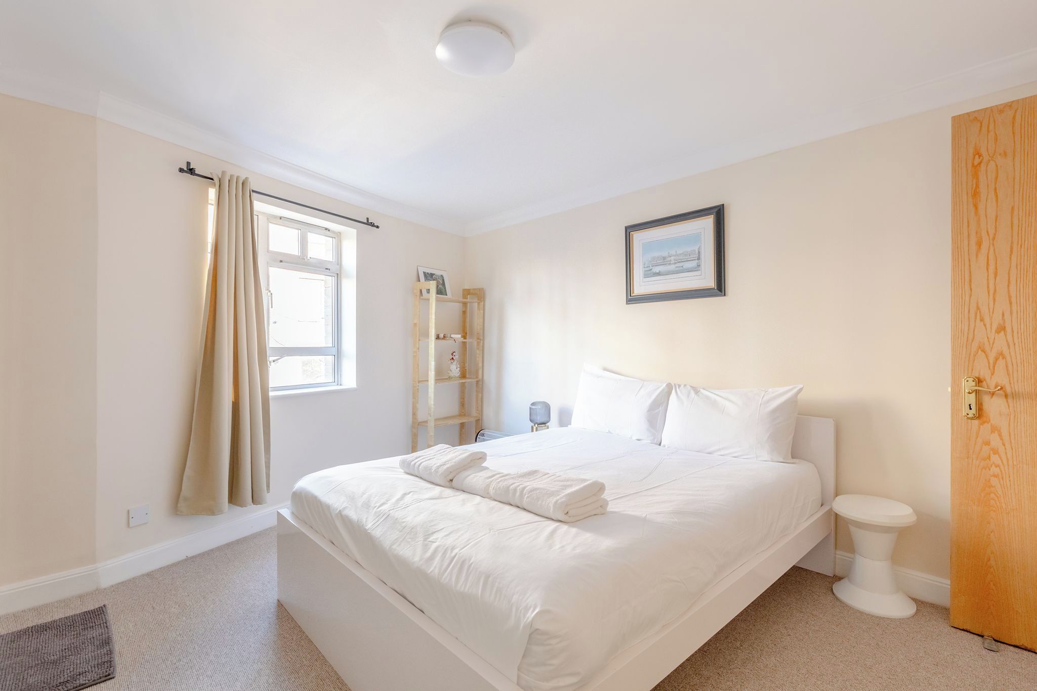 A bedroom with a bed, a window with curtains, a wooden door, and a picture on the wall.