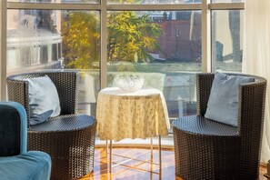 Een kamer met twee rieten stoelen, een klein tafeltje met een witte tafelkleed en uitzicht op gebouwen en bomen door grote ramen.