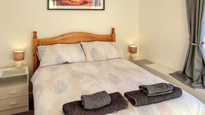 Ein ordentlich bezogenes Bett mit einem gemusterten Überwurf, zwei Nachttischlampen, einem hölzernen Kopfteil, einem kleinen Nachttisch und einem gerahmten Bild an der Wand.